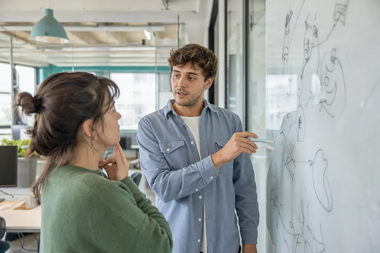 Junger Gründer erklärt einer Kollegin eine Skizze auf einem Glas-Whiteboard im hellen Co-Working-Space.